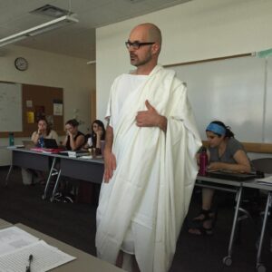 Matt Panciera standing in a toga. Behind him are several students seated at desks.